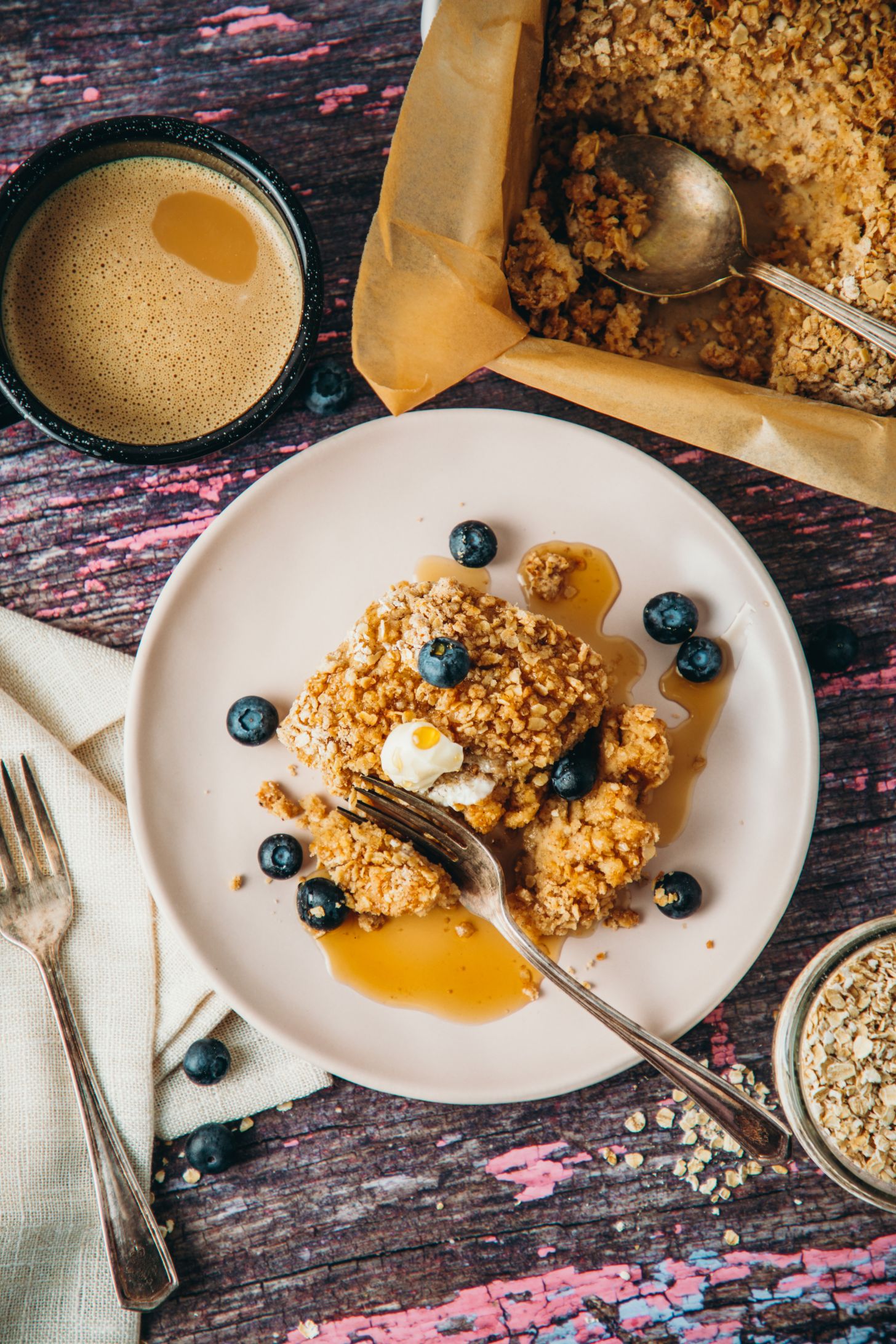 completed coffee cake pancake plated with blueberries and syrup as well as ingredients and utensils on the side against a dark background