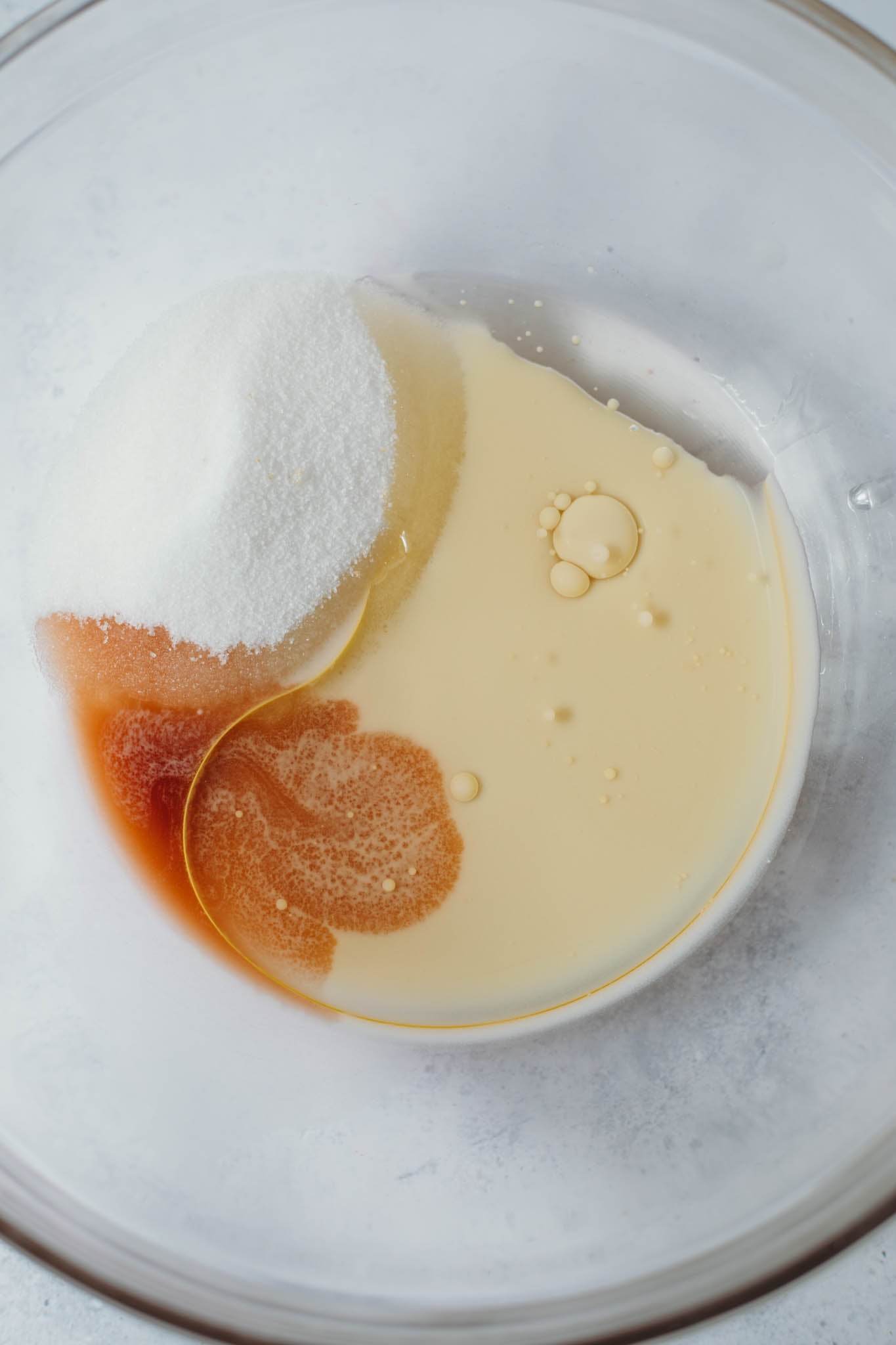sugar, vanilla and oil being mixed into the milk mixture from overhead on a white background.
