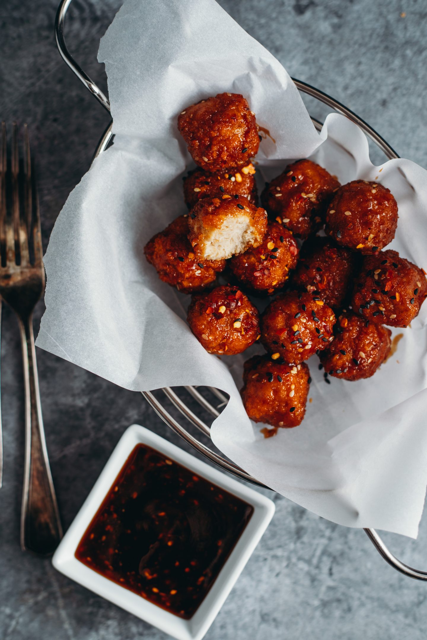 Delicious vegan teriyaki sauce drizzled over vegan chicken balls in a basket lined with white parchment paper. a fork and dish with dark teriyaki sauce to the side, and a bite has been taken out of one of the chicken balls. 