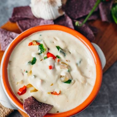 A white cheese sauce topped with spicy peppers in an orange clay dish styled with purple corn chips and a clove of garlic to the top left on a cutting board.